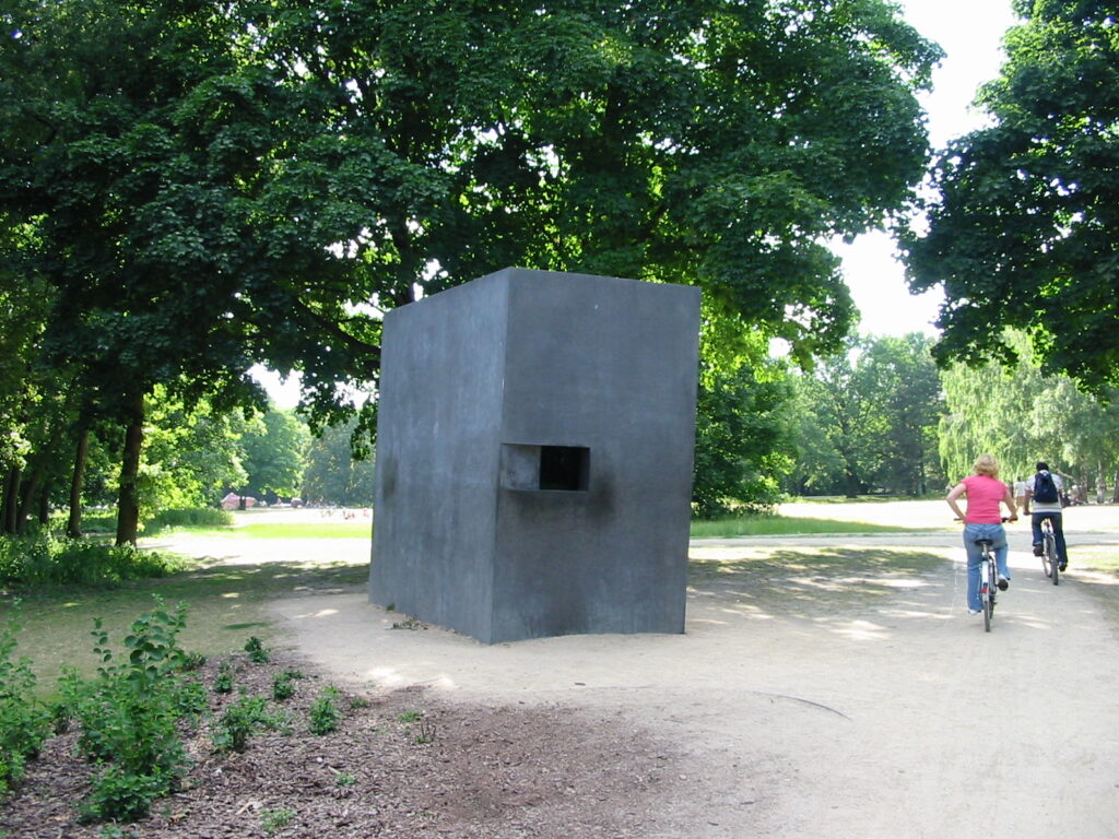 Et grå betonkasse, der er flere meter høj står i udkanten af en park. Der er et firkantet hul i øjenhøjde på en af kassens sider. Der er høje grønne træer omkring mindesmærket. To personer cykler forbi på en sti.